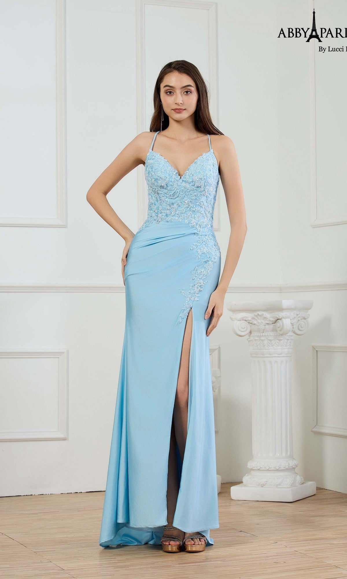 A woman stands indoors wearing the Abby Paris 90363 long prom dress in light blue, featuring beaded embroidery, spaghetti straps, lace accents, and a thigh-high slit.